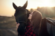 lovely young girl in a plaid shirt hugs a black horse at sunset in the mountains