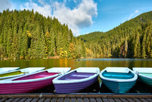 Multi-colored Boats At The Pier On Lake Lacu Rosu, Romania. Autumn Landscape Of Mountain Lake, Forest And Mountains.