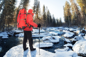 Hiker man with a red backpack stands on a stone by the river in a snowy forest among coniferous trees