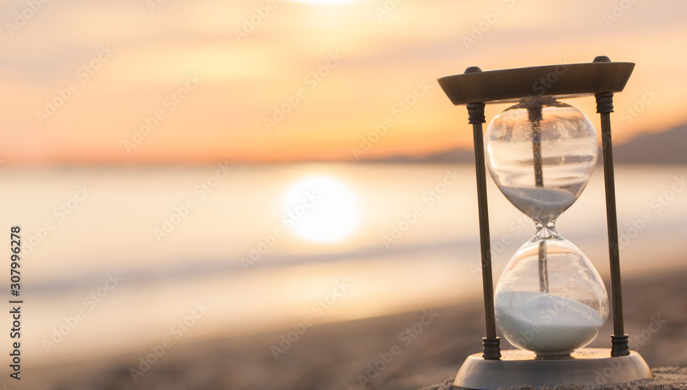Hourglass in the dawn time. Sand passing through the glass bulbs of an hourglass measuring the passing time as it counts down to a deadline or closure on a sunset/ sunrise beach background.  - obrazy, fototapety, plakaty 