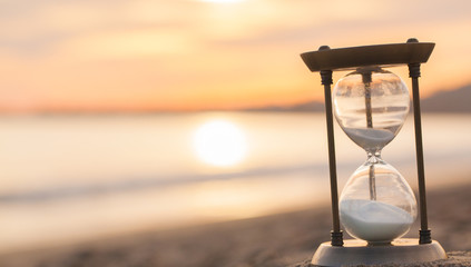 Poster - Hourglass in the dawn time. Sand passing through the glass bulbs of an hourglass measuring the passing time as it counts down to a deadline or closure on a sunset/ sunrise beach background. 