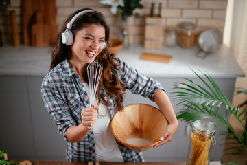 Wall Mural - Young woman in kitchen. Beautiful woman singing and dancing while cooking. Woman listening music while cooking.