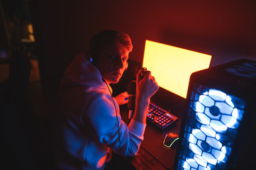 Wall Mural - Portrait of a serious young man in casual clothes sits at his computer at night in a dark room,holds abeer bottle in hand and looks int camera. Handsome gamer drinks beer, plays games on computer.