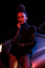 a young beautiful afro-american girl in a leather jacket, in a bodysuit posing on the hood of a car. stylish street light. night.