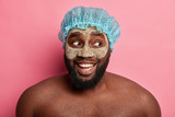 Fototapeta  - Curious happy bearded man looks aside, has thick bristle, wears clay mask, shower cap, waits for great result, looks aside, stands against rosy wall, enjoys procedure of cleaning face and skin