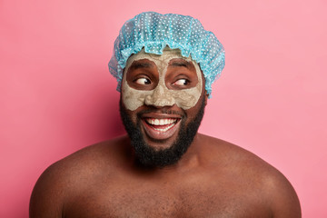 Curious happy bearded man looks aside, has thick bristle, wears clay mask, shower cap, waits for great result, looks aside, stands against rosy wall, enjoys procedure of cleaning face and skin