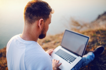 Rear view of handsome bearded blond caucasian man sitting on cliff and using laptop for internet surf.