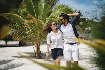 Wall Mural - Beautiful young couple joyfully walks on the white sand on the seashore between green palm trees. Honeymoon Travel and Vacation on the Tropical Ocean
