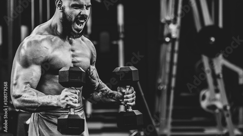 Brutal athletic bodybuilder training with heavy dumbbells in modern fitness club, dramatic black and white image