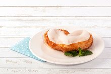 Homemade Glazed Puff Pastry In A Heart Shape With A Sprig Of Mint On A Porcelain Plate And Blue Polka Dot Paper Napkin. White Wooden Background