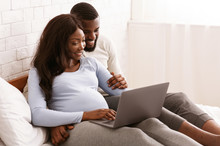 Pregnant Woman And Her Husband Using Laptop, Laying On Bed
