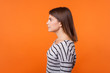 © khosrork - Side view portrait of calm confident young woman with brown hair in long sleeve striped shirt standing, looking focused and serious to the left. indoor studio shot isolated on orange background