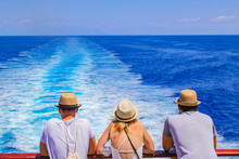 Tourists With A Straw Hat Stand On The Deck Of A Cruise Ship And Look Out Over The Ocean  While The Boat Is Sailing.