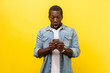 Portrait of amazed man in denim casual shirt using cellphone, typing text message or dialing number with shocked face expression, technology concept. indoor studio shot isolated on yellow background