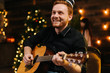 © dikushin - Laughing young man is playing guitar. Guy is looking happily and carefree. Man in festive hat celebrating Christmas or new year.