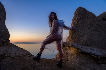 Silhouette of a young and sexy girl. She stands on a mountain and poses, dressed in a cloak and black boots against the sunset. Happy celebration victory success woman