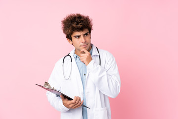 Young caucasian man over isolated pink background with doctor gown and holding a folder while thinking