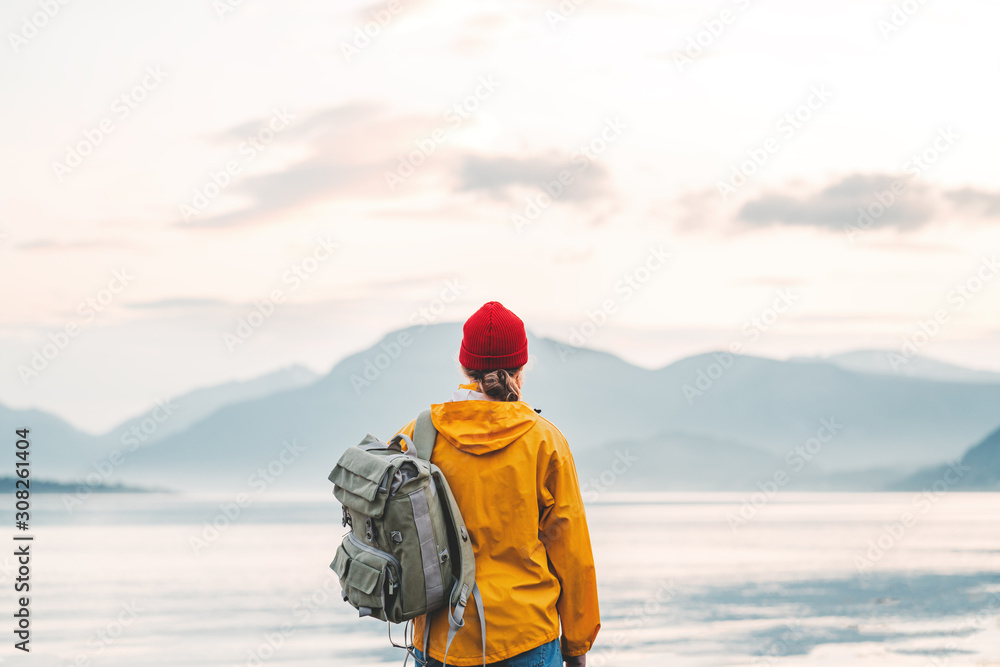Back view of male tourist with rucksack standing on coast in front of great mountain while journey.  Man traveler wearing yellow jacket with backpack explore scandinavia nature. Wanderlust outdoor - obrazy, fototapety, plakaty 