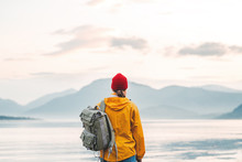 Back View Of Male Tourist With Rucksack Standing On Coast In Front Of Great Mountain While Journey.  Man Traveler Wearing Yellow Jacket With Backpack Explore Scandinavia Nature. Wanderlust Outdoor