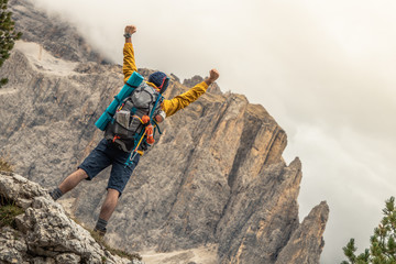 Wall Mural - young man hiker standing on stone mountain with raised up arms with cloudy sky and fog. yellow jacke