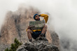 Young man hiker sitting on stone mountain reading map, with cloudy sky and fog. Yellow jacket, backpack, black beard and beanie. Traveling dolomites, Italy.