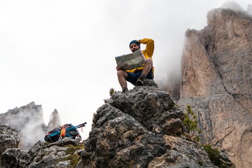 Wall Mural - young man hiker sitting on stone mountain reading map, with cloudy sky and fog. yellow jacket, backp