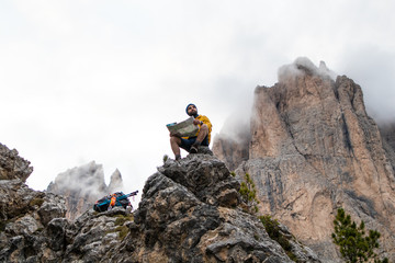 Wall Mural - young man hiker sitting on stone mountain reading map, with cloudy sky and fog. yellow jacket, backp