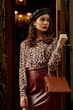© Victoria Fox - Outdoor fashion portrait of young elegant woman wearing black beret, leopard print blouse, faux leather dark red skirt, holding stylish brown bag, purse, posing in street of European city