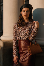 Outdoor Fashion Portrait Of Young Elegant Woman Wearing Black Beret, Leopard Print Blouse, Faux Leather Dark Red Skirt, Holding Stylish Brown Bag, Handbag, Posing In Street Of European City