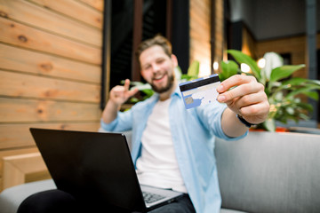 Wall Mural - Modern technology, business, travelling and online trading concept. Young Caucasian businessman holding credit card in one had and laptop on legs, making transaction. Focus on the card in hand