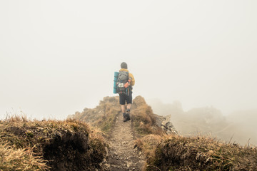 Wall Mural - young man hiker on mountain with cloudy sky and fog. yellow jacket, backpack and beanie. traveling d