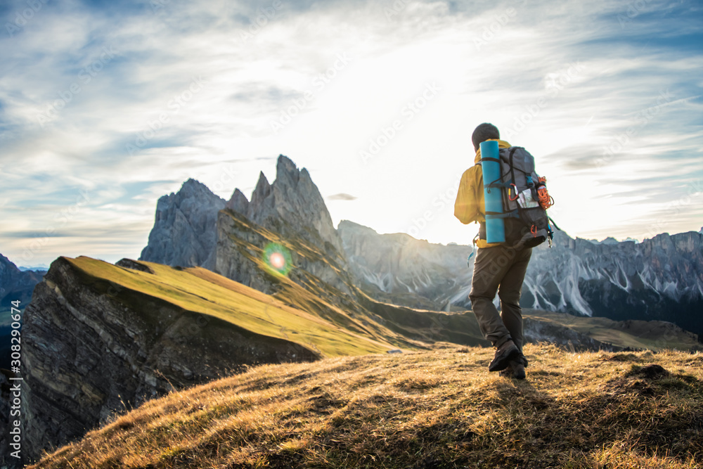 Young man hiking at Seceda mountain peak at sunrise. Backpack, yellow jacket, boots, beanie. Traveling to puez Odle, Dolomites, Trentino, Italy. - obrazy, fototapety, plakaty 