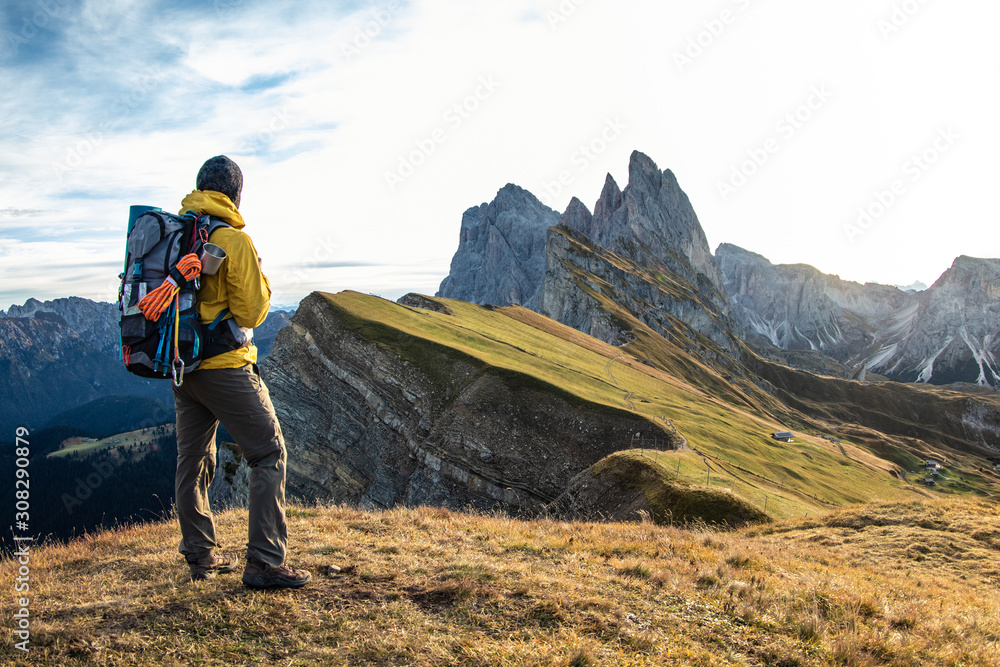 Young man hiking at Seceda mountain peak at sunrise. Backpack, yellow jacket, boots, beanie. Traveling to puez Odle, Dolomites, Trentino, Italy. - obrazy, fototapety, plakaty 