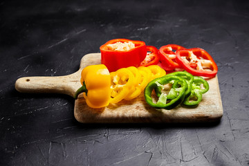 Cutting board with slices of red, green, and yellow bell peppers on black background. Sliced sweet peppers in different colors on stone table, vegetable ingredient, cooking healthy food