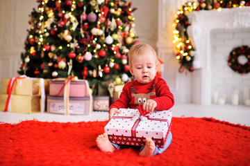 a child-a little boy in a red Christmas sweater with a picture of Santa's reindeer sits barefoot on a warm red carpet with a gift in his hands on the background of a Christmas tree and decor