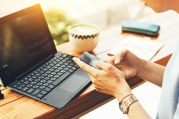 Close-up of female hands holding a mobile phone and writing sms, in the background a laptop and a cup of tea on the table by the window in a cafe