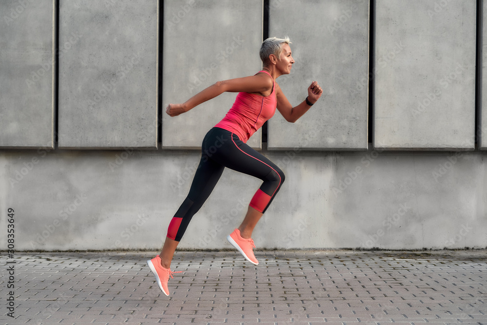 Full of energy. Side view of active middle aged woman in sport clothing jumping while exercising outdoors - obrazy, fototapety, plakaty 