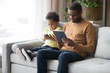 © fizkes - Black father and little son use gadgets sitting on couch