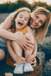 Portrait of a lovely little girl laughing with closed eyes while is embraced by her mother with is laughing and looking at camera against sunset.