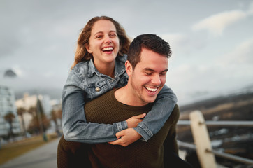 Canvas Print - Happy young friends dating outdoor in city sunset, man carrying a woman on his back