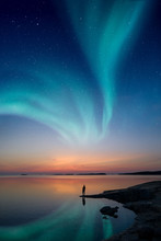 A Man Standing By A Calm Water And Looking At The Northern Lights On The Sky With Reflections From The Water