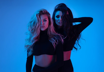 Blonde and brunette models in black clothes looking at camera and posing in studio on blue background 