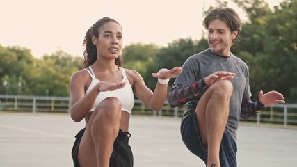 Wall Mural - Sporty young couple in sportswear doing exercise on their legs and smiling while working out at playground outdoors