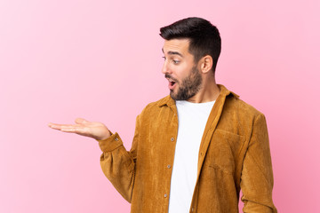 Wall Mural - Young handsome man with beard wearing a corduroy jacket over pink background holding copyspace imaginary on the palm