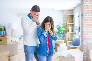 Poster - Young beautiful couple standing at new home around cardboard boxes with sad expression covering face with hands while crying. Depression concept.