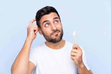 Young handsome man with beard brushing his teeth over isolated background having doubts and with confuse face expression