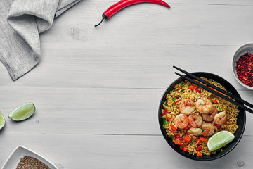 Wall Mural - Shrimp fried rice with vegetables, green onions and lime in a black pottery bowl. Traditional Thai meal on a white table with fabric tablecloth. Top view, directly above shot with space for text.