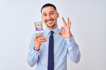 Poster - Young handsome business man holding a bunch of dollars bank notes over isolated background doing ok sign with fingers, excellent symbol