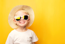 Portrait Of A Cute Blond Baby Boy With A Happy Face Smile In A Straw Hat And Sunglasses On A Yellow Background Looking At The Camera. Card With Place For Text
