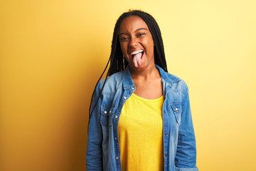 Young african american woman wearing denim shirt standing over isolated yellow background sticking tongue out happy with funny expression. Emotion concept.